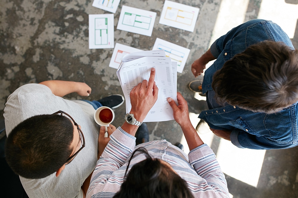Top view of hands of three young people discussing financial transactions during a meeting. Man pointing at document while standing with colleagues in office. Business partners discussing financial data at meeting