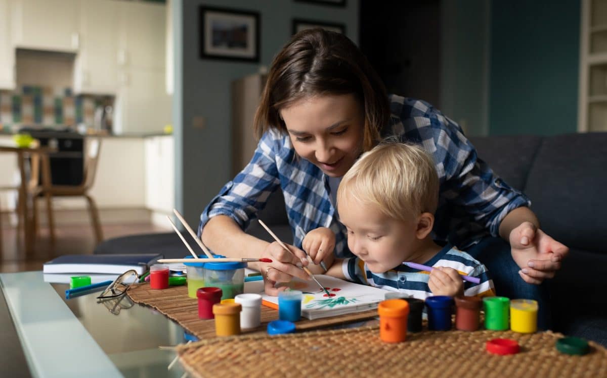 Positive female babysitter in casual clothing helping with painting to small blond haired boy sitting on couch at living room at home